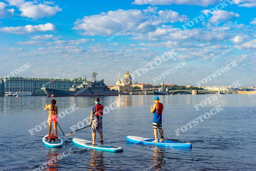 На фотографии группа людей занимается сапсерфингом на реке Неве в Санкт-Петербурге. На заднем плане видны Эрмитаж, Исаакиевский собор и военный корабль, создающий контраст между исторической архитектурой и современностью. Голубое небо и легкие облака добавляют ощущение легкости и радости летнего дня.